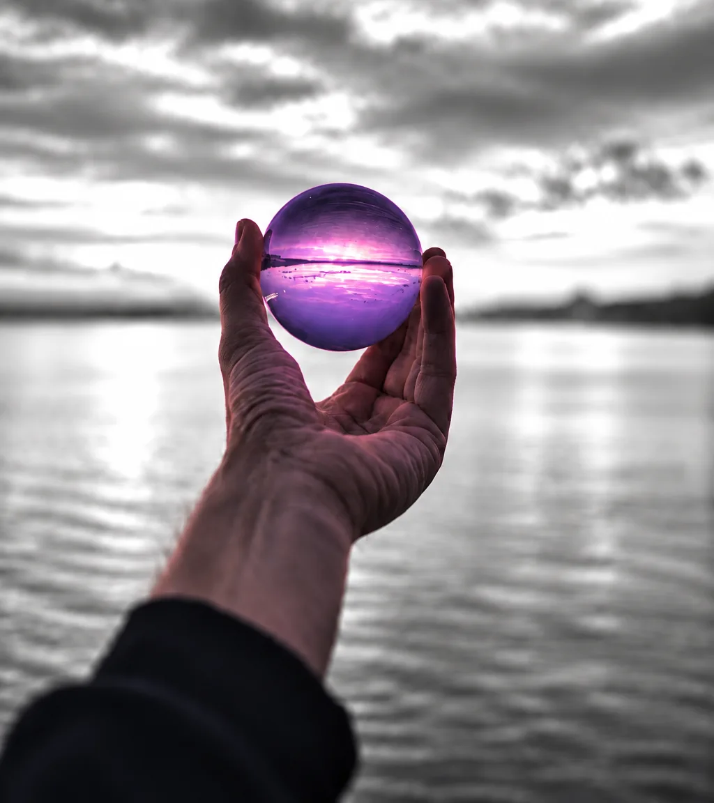 Hand hält eine violette Glaskugel vor einem grauen Wolkenhimmel über dem Wasser